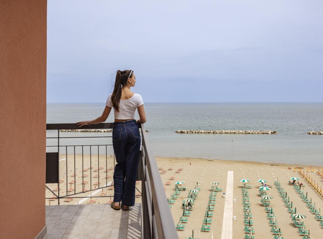 Woman on balcony gazes at sea, empty beach, peaceful atmosphere.